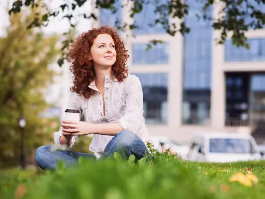 Rothaarige Frau sitzt mit To-Go-Kaffeebecher im Park, im Hintergrund ein Bürogebäude 