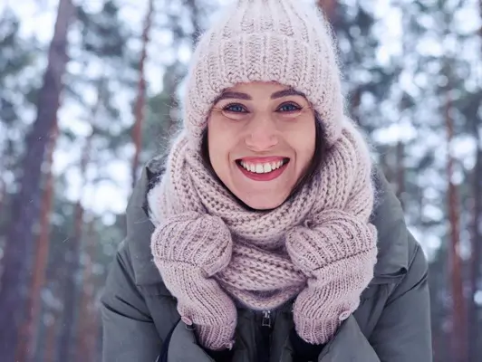Junge Frau in Schal und Mütze lächelt vor winterlicher Landschaft in die Kamera