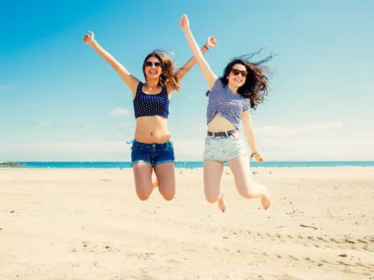 Zwei hübsche Frauen in Jeans-Shorts und schlanken Beinen springen fröhlich an einem Strand herum