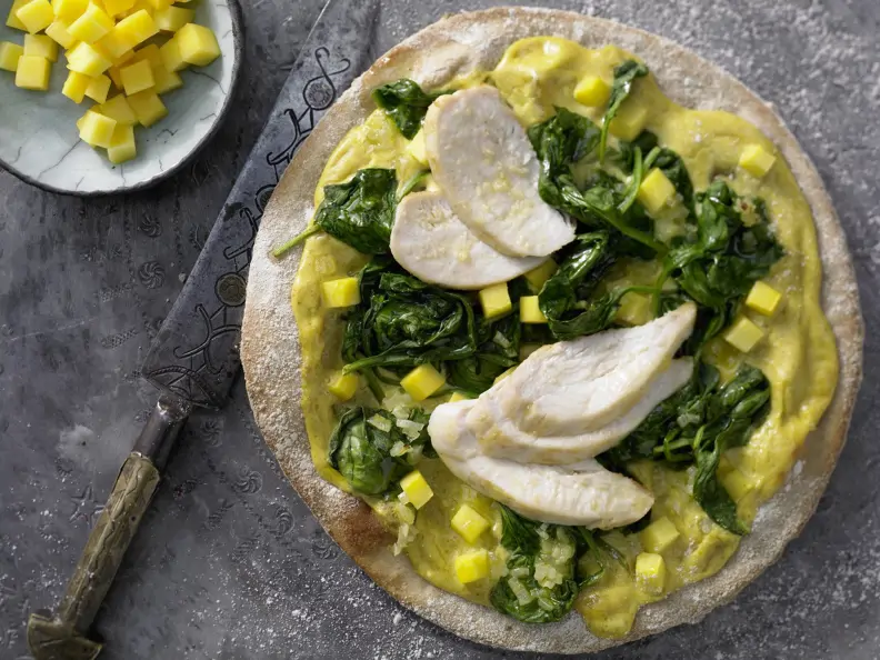 Pizza auf indische Art mit Hähnchen auf einem grauen Untergrund, daneben eine Schale mit Mangowürfel