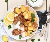 Paniertes Lachsfilet auf hellem Teller und Zeitungspapier, dazu selbstgemachte Kartoffelchips und Dip