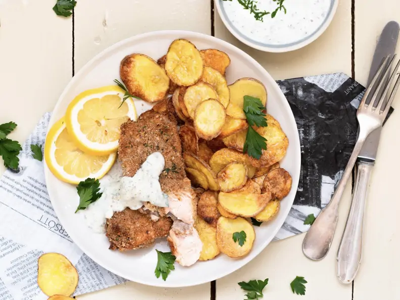 Paniertes Lachsfilet auf hellem Teller und Zeitungspapier, dazu selbstgemachte Kartoffelchips und Dip