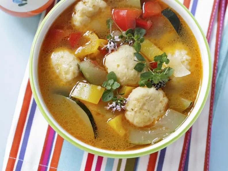 Gemüsesuppe mit Käsebällchen in weiß-grüner Schale auf buntem Set