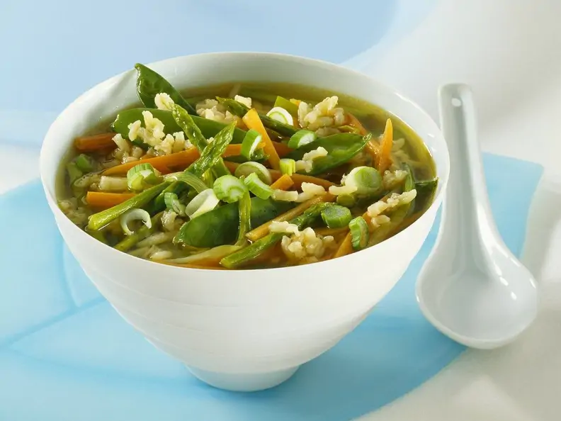 Gemüsesuppe mit Spargel und Reis in Schale neben weißem Löffel